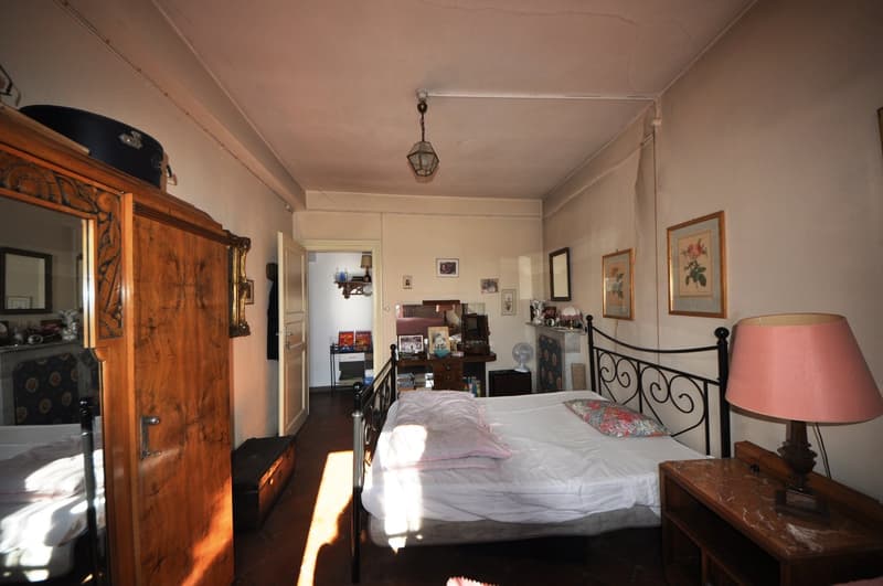Spacious bedroom with a wooden armoire, a metal bed frame, a nightstand, and various decorative items and artwork on the walls. The room has a cozy and eclectic feel.