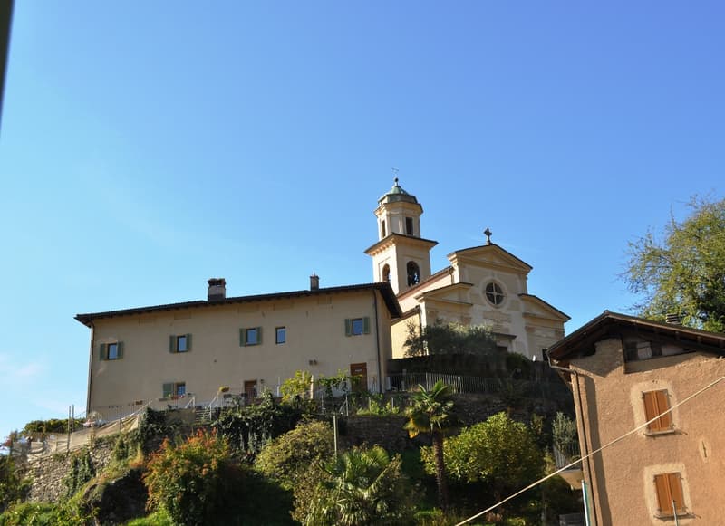 A multi-story building with a church-like tower, surrounded by lush greenery and vegetation. The building has a Mediterranean architectural style with a domed roof and arched windows.