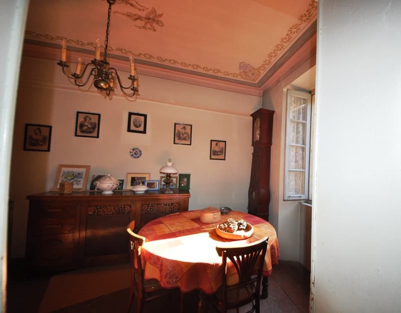 The image shows a cozy and traditional dining room with a chandelier, ornate ceiling, framed artwork on the walls, and a wooden cabinet or sideboard. The dining table is set with a patterned tablecloth and a decorative centerpiece. The room has an overall