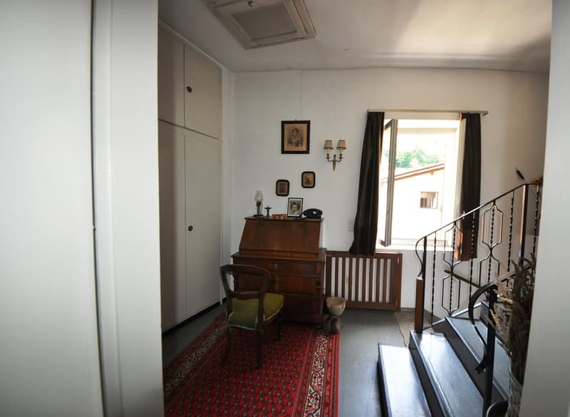 The image shows a living room with a wooden desk, framed artwork on the walls, and a fireplace. The room has a traditional, vintage decor with a red patterned rug on the floor.