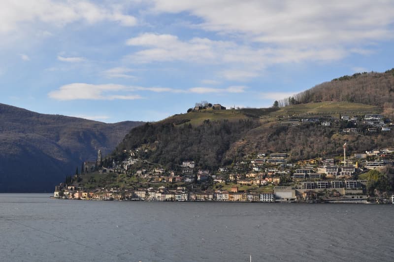 The image shows a scenic view of a town nestled in the hills along a body of water, likely a lake or river. The town features a mix of residential and commercial buildings, with some larger structures visible on the hilltops. The landscape is dominated by