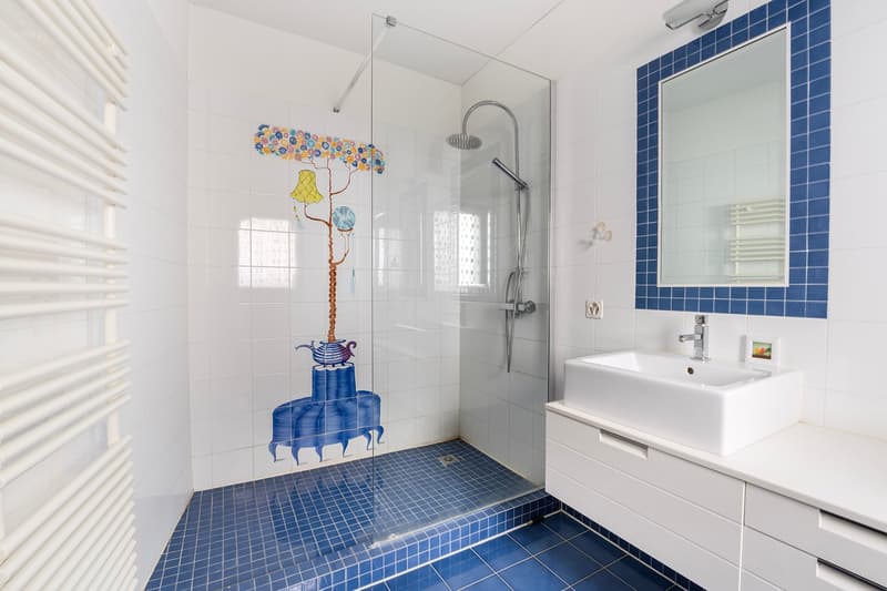 Spacious bathroom with white tiles, blue accent tiles, a walk-in shower, a white vanity with a sink, and a decorative lamp fixture with colorful elements.