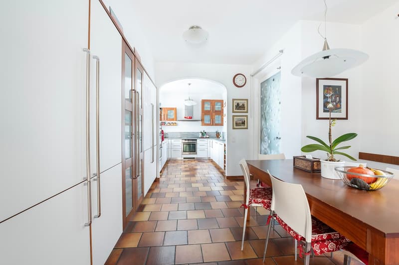 Spacious kitchen with white cabinets, tiled floor, and stainless steel appliances including a dishwasher. The kitchen opens up to a dining area with a wooden table and chairs. The space has a bright and airy feel with large windows and decorative wall art