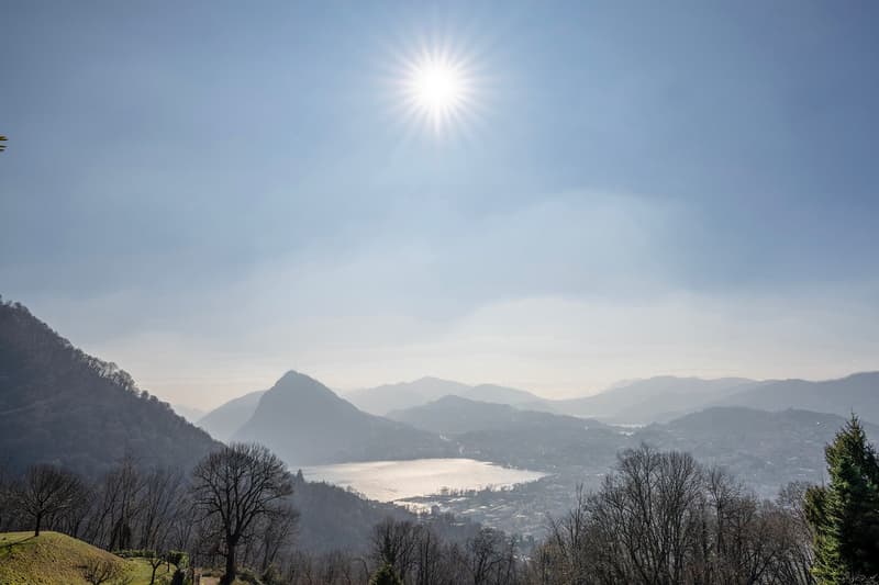 The image shows a scenic mountain landscape with a lake in the foreground. The sun is shining brightly in the sky, casting a warm glow over the scene. The mountains in the distance are partially obscured by haze, creating a sense of depth and atmosphere.