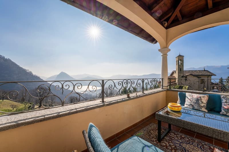 Spacious balcony with ornate wrought-iron railing, offering a panoramic view of the surrounding mountains and a church steeple in the distance. The balcony is furnished with wicker furniture and decorative pillows, creating a cozy and relaxing outdoor spa