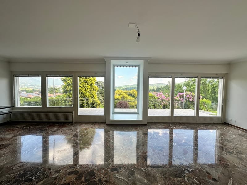 Spacious living room with large windows offering a scenic view of the surrounding landscape, including trees and flowers. The room has a marble floor with a reflective surface, creating a luxurious and elegant atmosphere.
