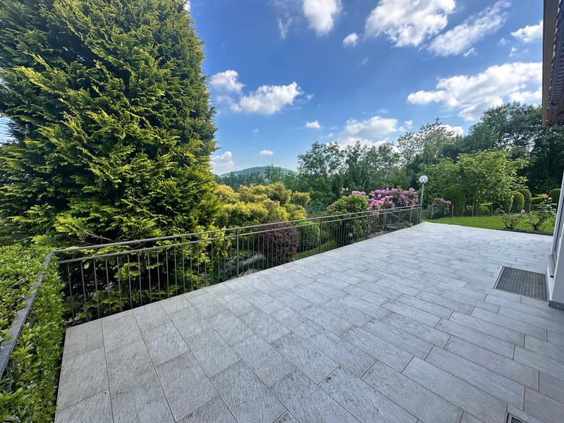 The image shows a spacious outdoor patio or deck area with a stone tile floor. The patio is surrounded by lush greenery, including tall evergreen trees and flowering shrubs. There is a metal railing along the edge of the patio, providing a balcony or terr