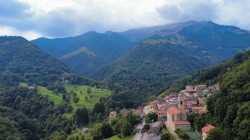 village with stone houses, red roofs, hilly terrain, dense greenery, terraced rice fields, mountain range, cloudy sky