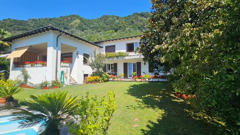 Two-story house, white exterior, awnings, green plants, red flowers in planters, swimming pool, expansive garden, mountainous background