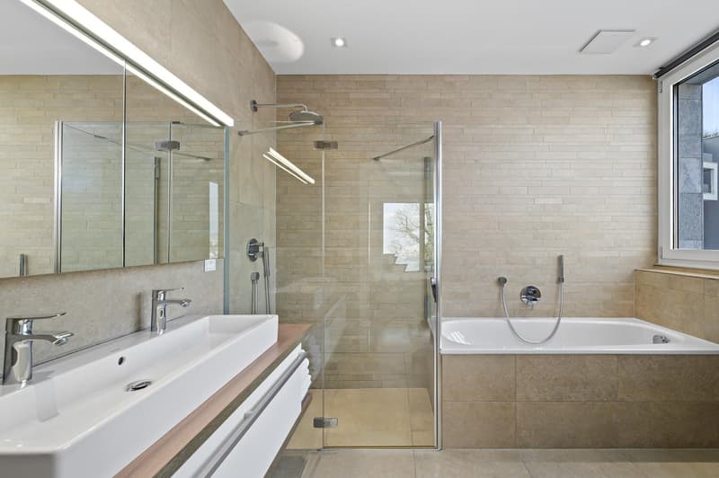 Modern bathroom with glass shower enclosure, white bathtub, beige tiles, large mirror, white sink, faucets, towel rack, built-in lighting.