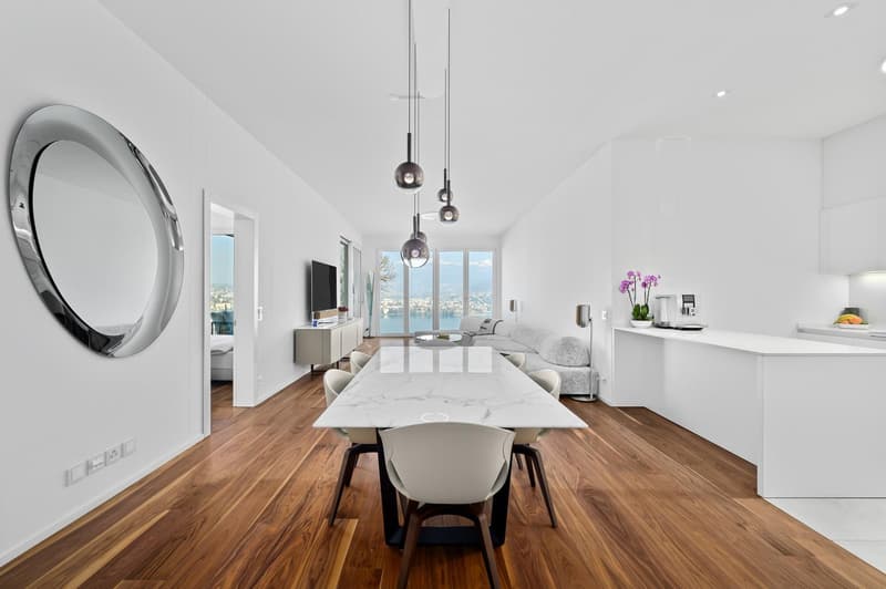 Modern interior with hardwood floors, a large marble dining table with chairs, a white marble kitchen island, a wall-mounted television, a couch with a center table, a decorative plant, and a round mirror.