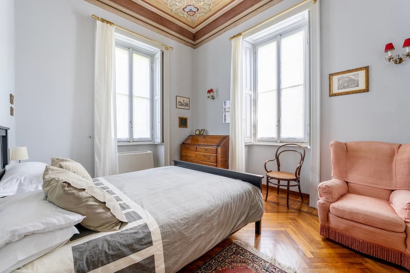 Bedroom with bed, pillows, two windows, wooden floor, bedside table, lamp, armchair, desk, wooden cabinet