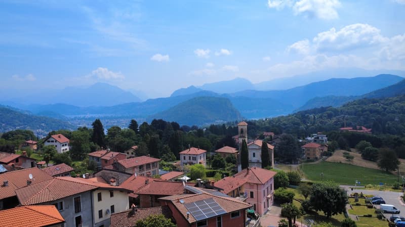 Aerial view of a village with tiled roofs and lush greenery, surrounded by mountains and a lake, featuring a church and a tower.