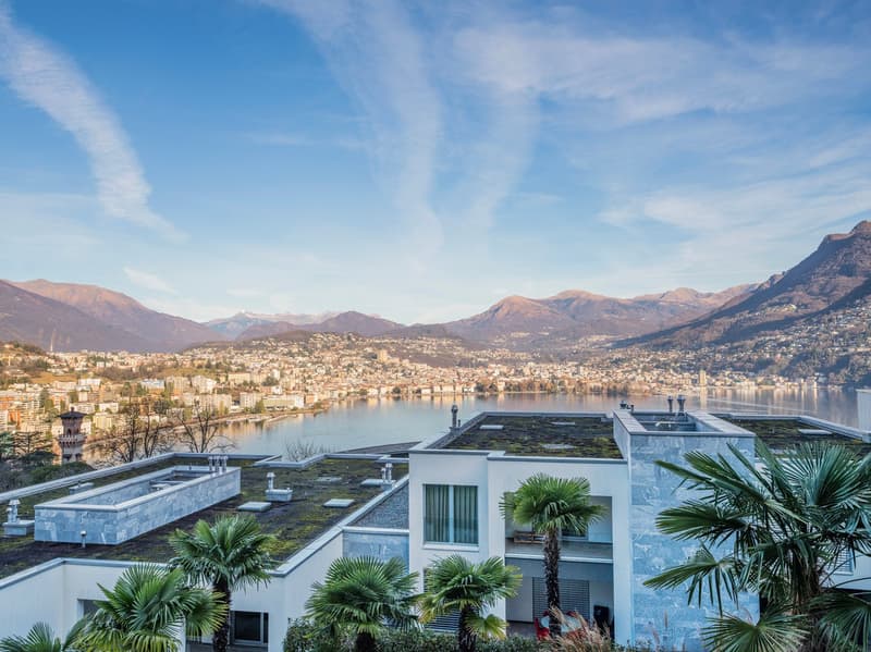Aerial view of a white building on a hill with lush greenery, surrounded by mountains, a lake, and a cityscape in the background