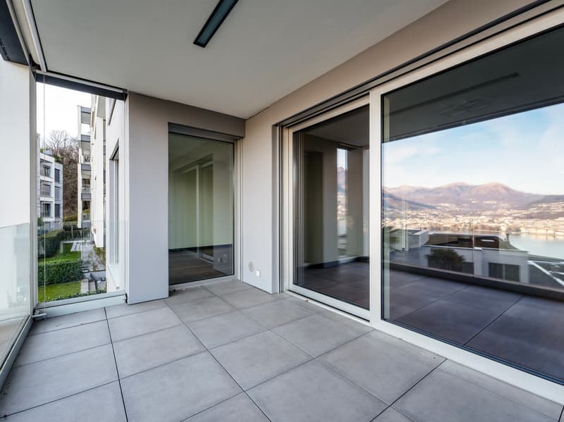 Empty balcony with tile floor, glass walls and doors, overlooking a landscaped area and a lake with mountains
