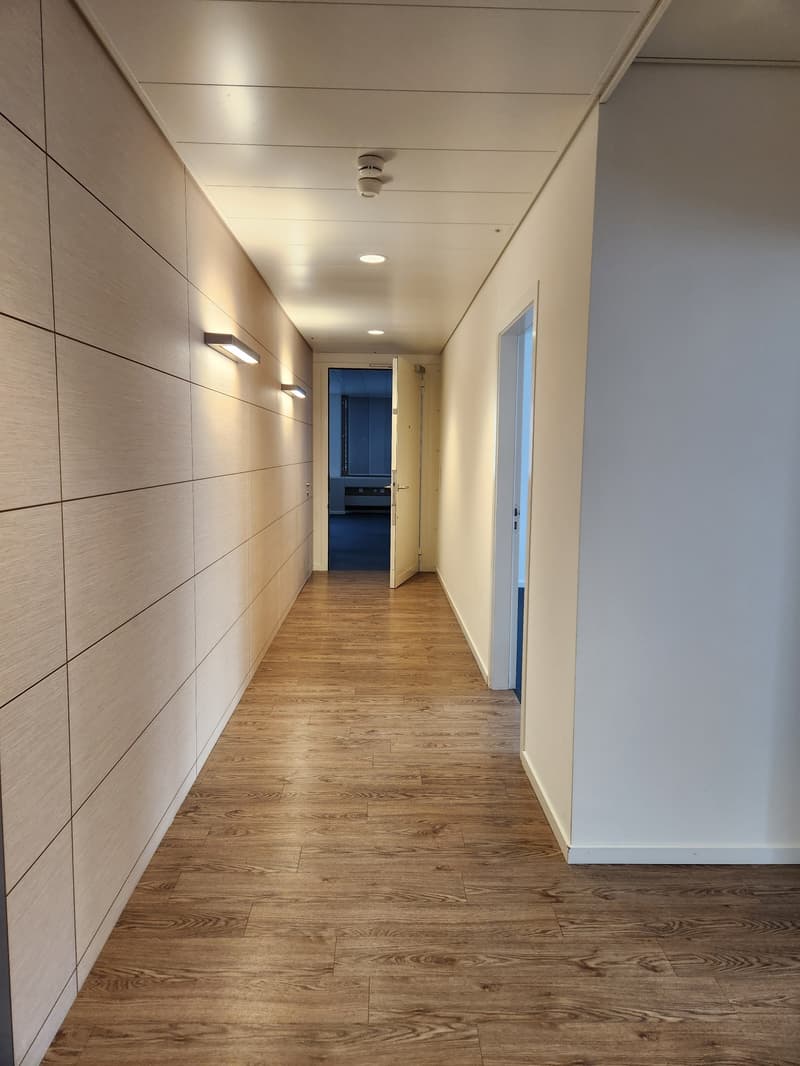 Empty corridor with wooden floor and beige walls, two open doors, one to a room and one to a larger room