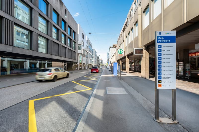 The image shows a city street in Zug, Switzerland. The street has multiple buildings on both sides, with a mix of commercial and residential properties. There are yellow lines on the road indicating parking spaces, and a sign post with information about t