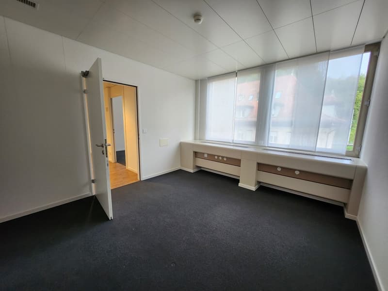 Large empty room, white walls, carpeted floor, large windows with curtains, open door leading to another room