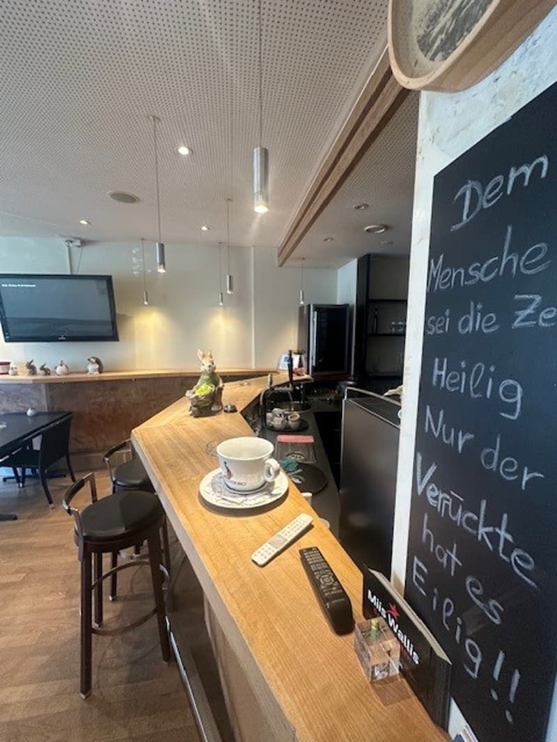 A counter with a cup and saucer, remotes, and a blackboard with writing. Behind the counter, there is a sink with a faucet, a refrigerator, and a television. There are chairs and a table in the room.