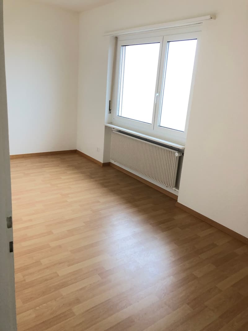 The image shows a spacious bedroom with white walls and hardwood flooring. The room has two large windows that provide ample natural light. There is a radiator installed under the windows.