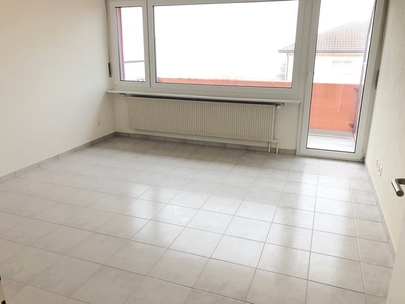 The image shows a spacious room with large windows, a tiled floor, and a radiator along the wall. The room appears to be empty, with no furniture or decor visible.
