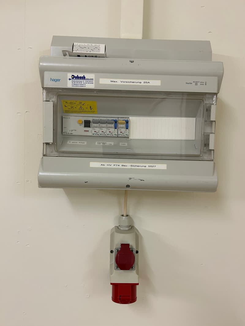 Gray metal circuit breaker box, multiple fuses, labels with voltage and circuit ratings, red toggle switch, plug socket with plug connected