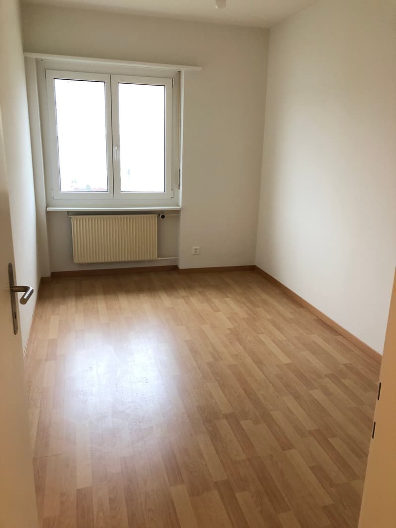 Empty room with wooden floors, white walls, a window, a radiator, and power outlets