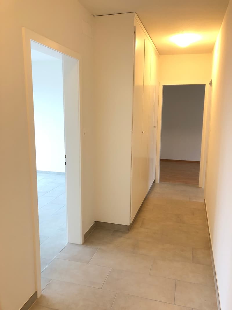 Corridor with tiled floor, white walls, two doorways, a ceiling light, and a wooden floor in the right room.