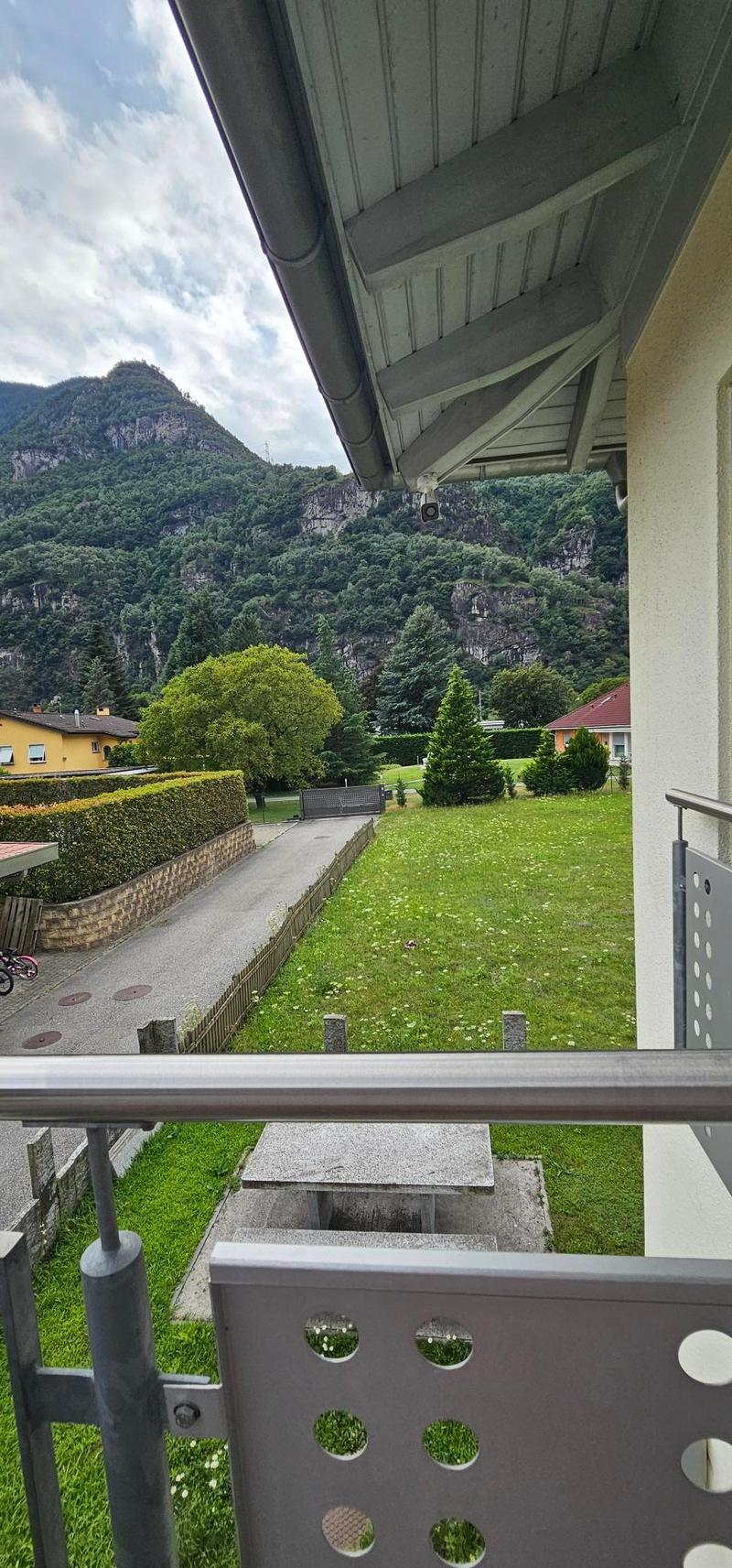 The image shows a balcony or terrace overlooking a scenic landscape. The balcony has a wooden structure and railing, and the view includes a grassy area with trees and shrubs, as well as a mountainous backdrop in the distance.