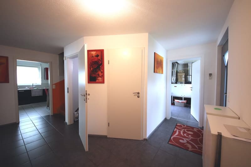 Modern hallway with tiled floor, open door, white walls, framed artwork, a rug, cabinets, and a kitchen area in the background.
