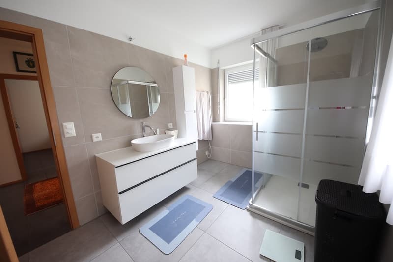 Modern bathroom with white tiled walls, glass shower enclosure, vanity with mirror, white vanity, blue mats, and towels hanging from the rack.