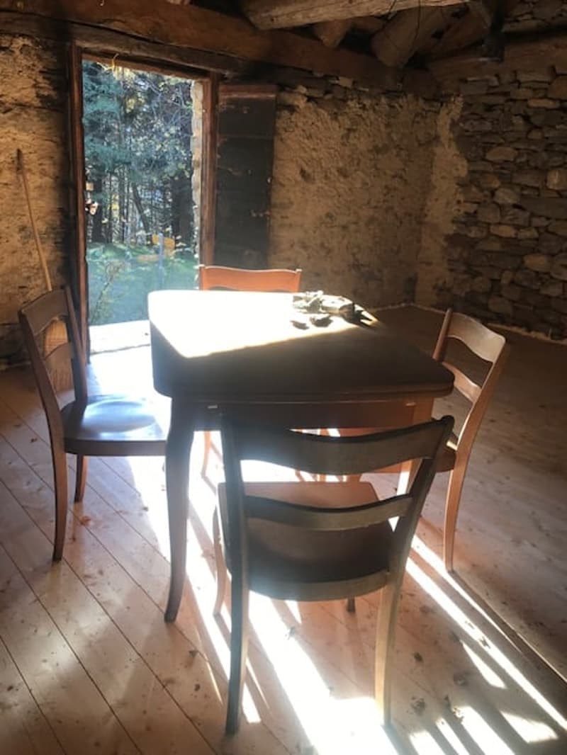 Rustic dining room with stone walls, wooden beams, and a wooden dining table with chairs. The room has a large window that provides a view of the outdoor area.