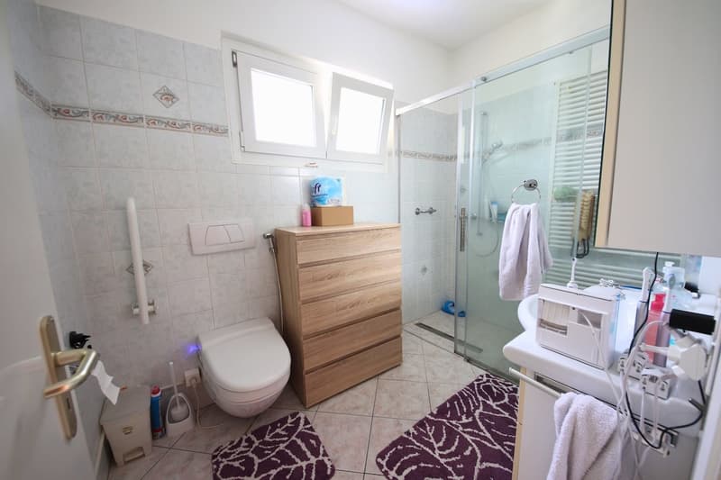 White tiles, white toilet, glass shower enclosure, wooden cabinet, sink with countertop, towels, and various bathroom items
