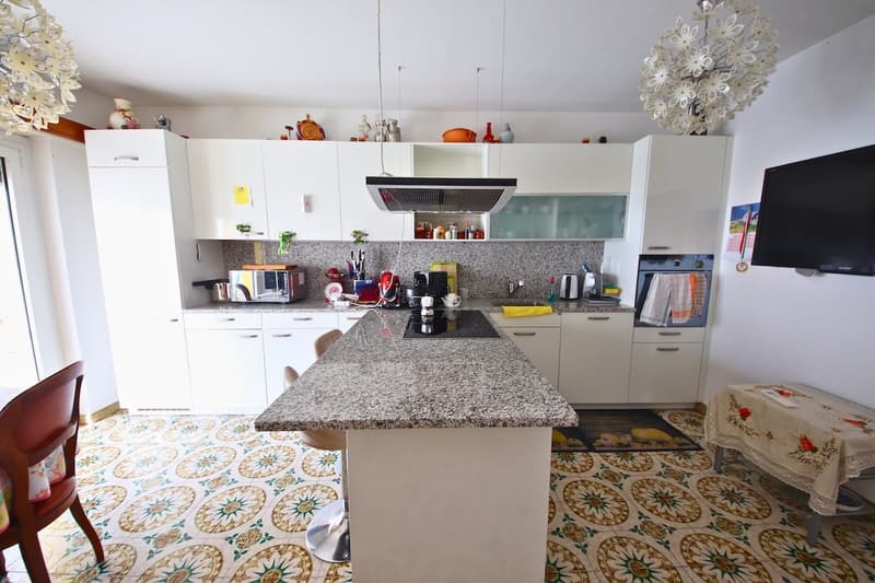 White cabinetry, granite counter, central island with 2 stools, built-in oven, microwave, refrigerator, induction cooktop, dishwasher, kitchen sink, modern appliances, decorative items on shelves