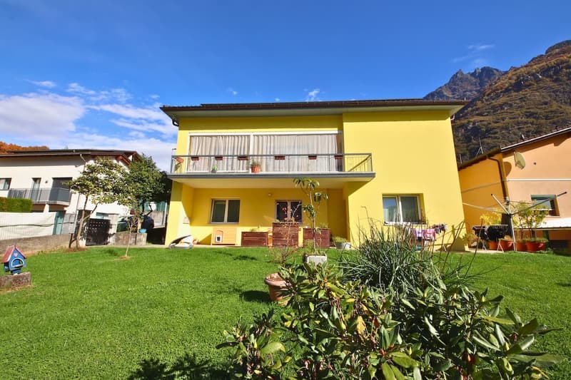 2-story house, yellow paint, white windows, terrace with metal railing, front yard with green grass, mountain in the background