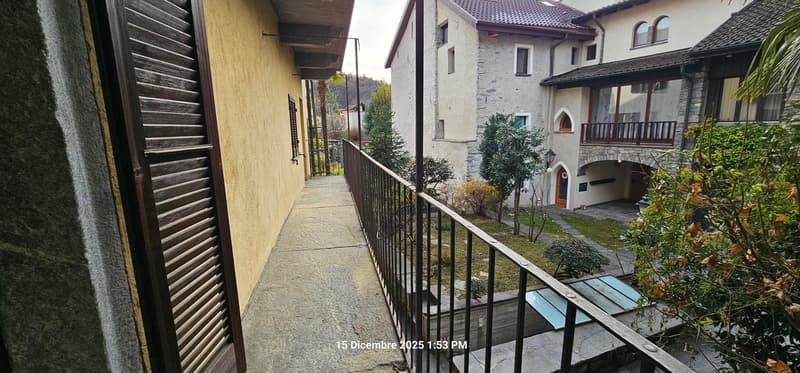 Building with balcony, stone walls, plants, pathway, trees, and cars parked in the distance