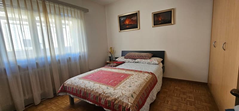 single bed, wooden floor, white walls, floral blanket, nightstand with flower vase, two paintings on wall, curtained window, wooden closet