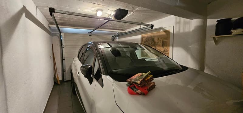 Garage: White car, ceiling lights, wooden storage box, white tiled floor, picture frame on the wall, and helmets on the shelf