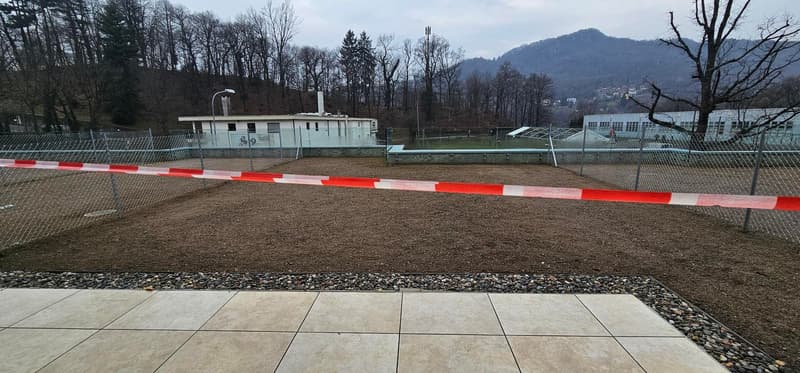Fenced area with soil, surrounded by buildings, no vegetation, streetlights, mountains in the distance