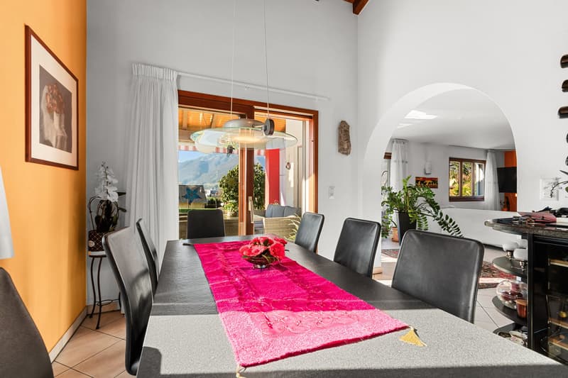 Dining room with a large table, chairs, a pink table runner, a flower vase, a picture frame, and a glass door leading to a balcony with a view of the mountains.