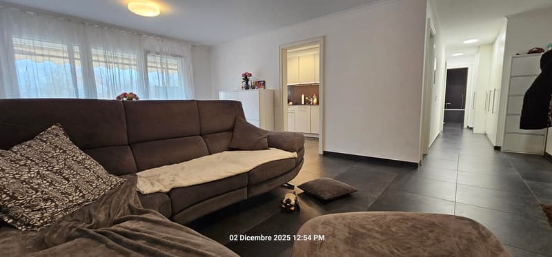 A room with modern L-shaped couch, white walls, black tiled floor, and natural light coming through large windows with curtains.