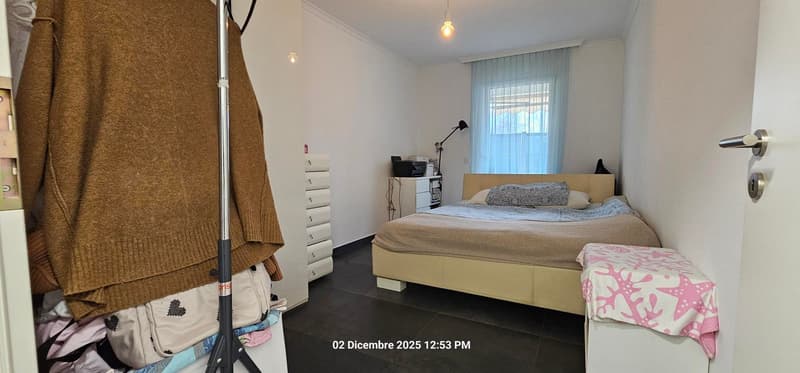 A bedroom with white walls, black tiled floor, a bed, a cabinet, a lamp, and a window with curtains.
