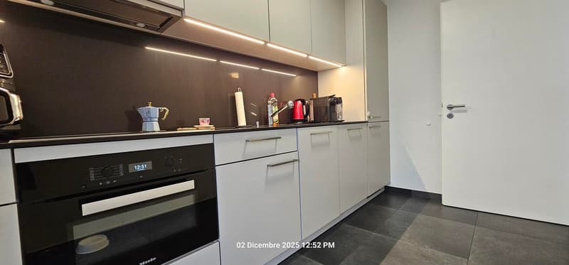 Kitchen with oven, sink, countertop, cabinets, drawers, microwave, kettle, coffee maker, coffee cups, plates, bottle, roll of paper towels, clock, watermark