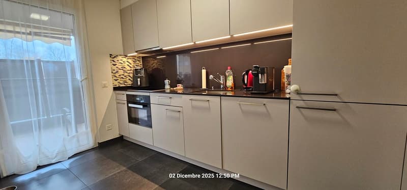 modern kitchen with white cabinets, black countertops, built-in oven and microwave, sink with faucet, kitchen appliances including a coffee machine, toaster, kettle, blender, and various utensils and bowls on the countertop.