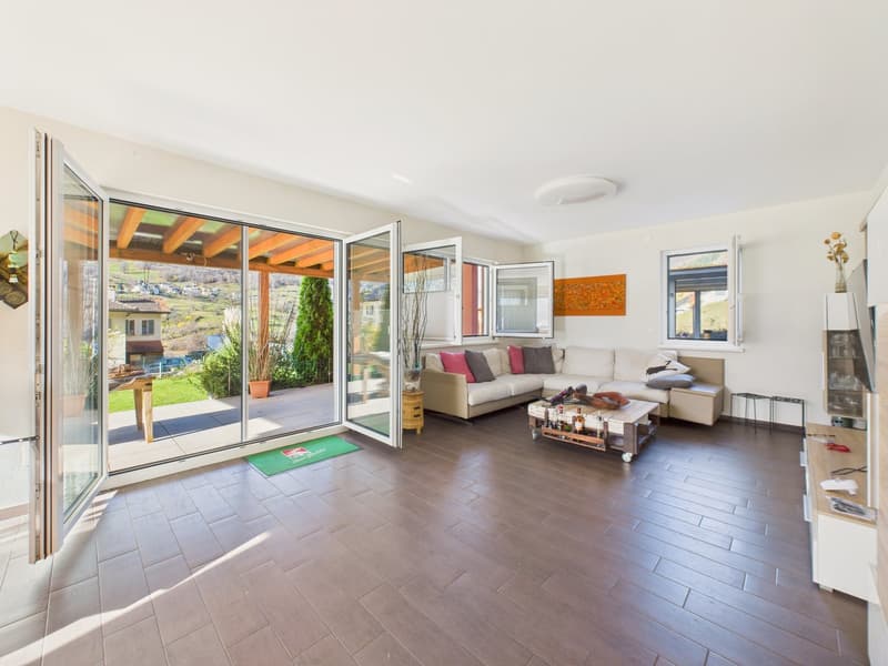 Open living room with large glass doors leading to a terrace, wooden floor, white walls, beige sofa, coffee table, and a view of the landscape outside