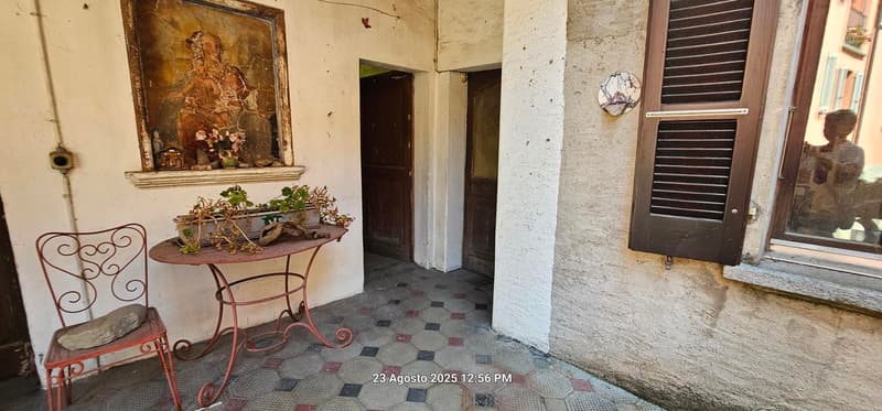A narrow corridor with a tiled floor, small table with a plant, a chair, a wall with a framed painting, closed door and a window with wooden shutters