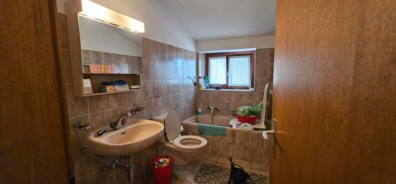 bathroom with toilet, bathtub, sink, mirror, brown tiled walls, small wooden shelf with soap, small window, light above mirror