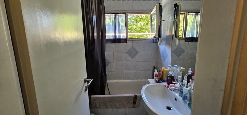 Bathroom with bathtub, sink, and shower curtain. Tiled walls and floor. Various toiletries on the sink.