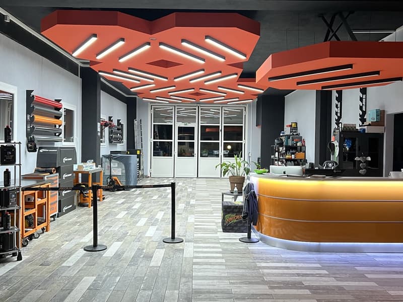 Commercial shop with modern and minimalistic design, orange and white color scheme, LED lighting, wooden floor, reception desk, tables, storage units, and a small potted plant.
