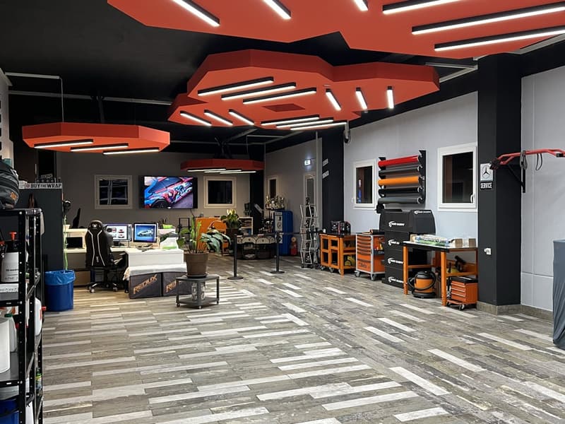 Interior of a car wrap shop, featuring red octagonal ceiling lights, large tables with computers, storage units, and tools, a monitor displaying an image, office chairs, and shelves with items.